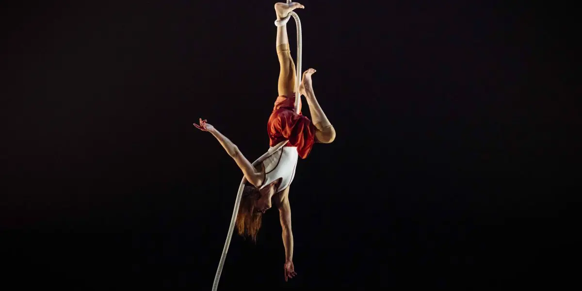 L'artiste aérienne Rosaleen Rogmans est suspendue à une corde, les jambes entrelacées, les bras gracieusement étendus, vêtue d'un haut blanc et d'un short rouge, sur un fond sombre.