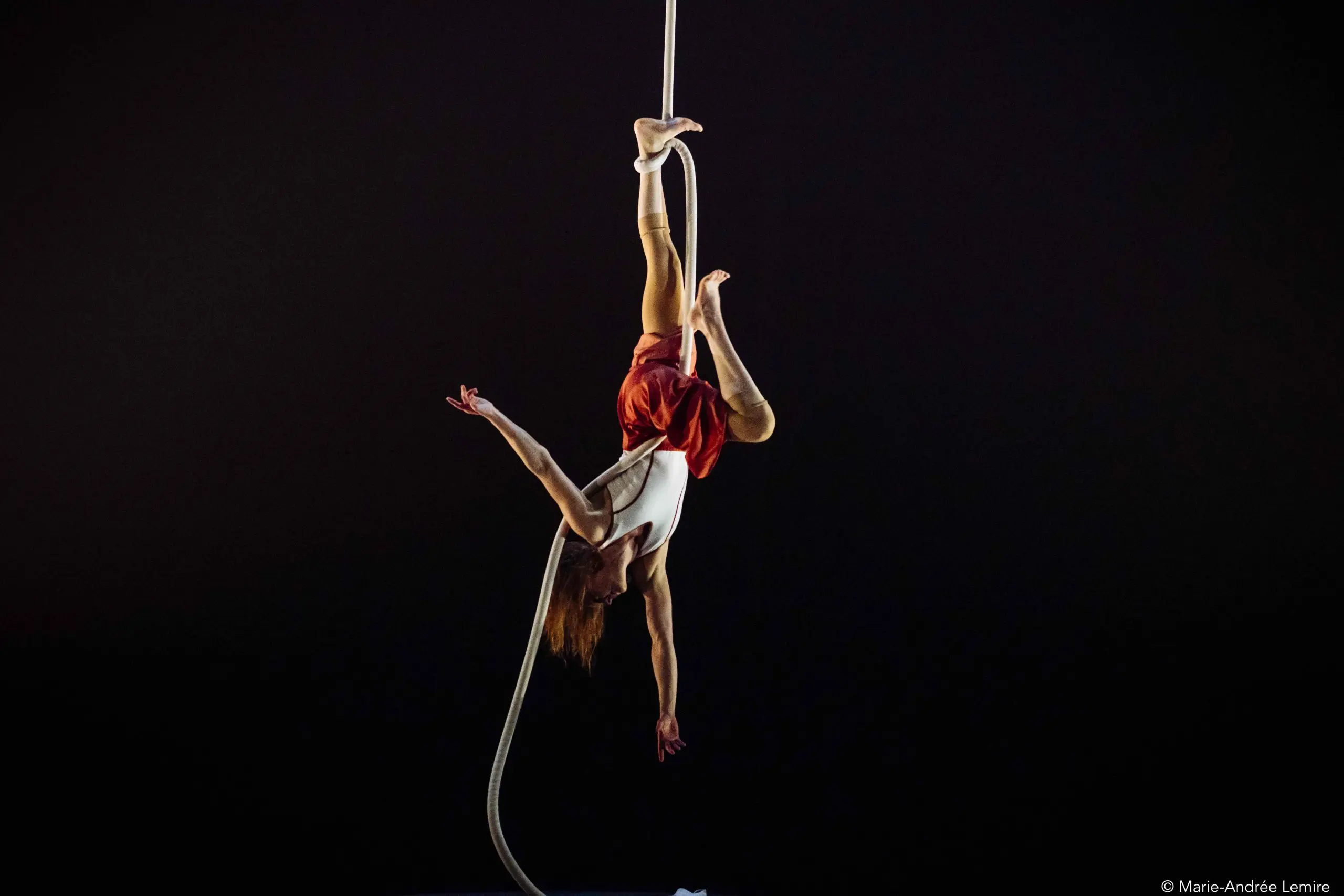 L'artiste aérienne Rosaleen Rogmans est suspendue à une corde, les jambes entrelacées, les bras gracieusement étendus, vêtue d'un haut blanc et d'un short rouge, sur un fond sombre.