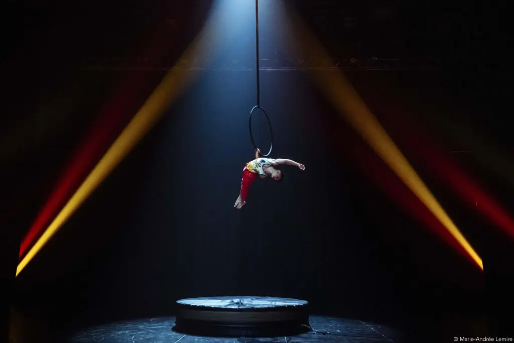 Un acrobate, Enzo Pebre, se produit sur un cerceau aérien suspendu sous un projecteur sur une scène sombre, avec deux faisceaux de lumière jaune et rouge qui brillent en diagonale à l'arrière-plan.