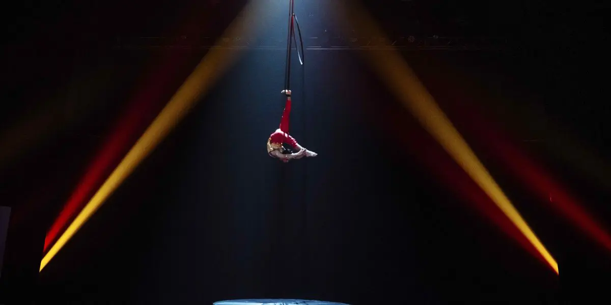 Enzo Pebre, un artiste aérien vêtu d'un costume rouge, est suspendu à un cerceau à l'envers sur une scène sombre, éclairée par un projecteur dramatique et flanquée de faisceaux de lumière orange et jaune.