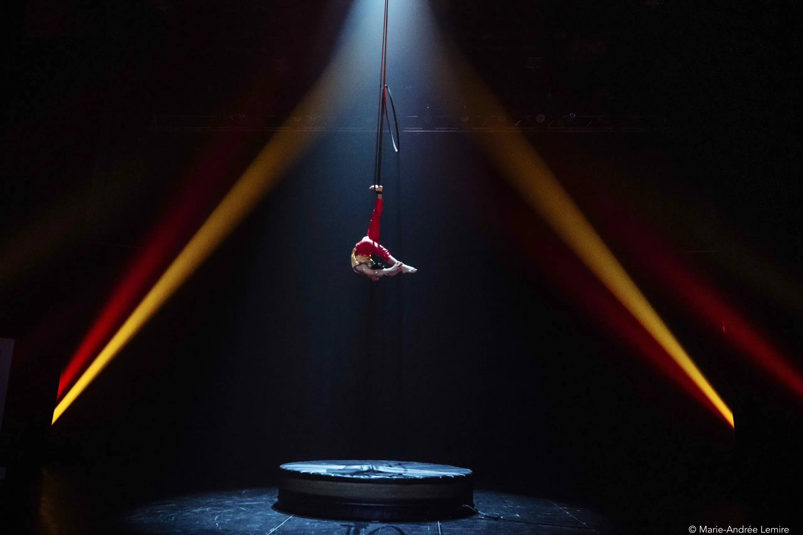 Enzo Pebre, un artiste aérien vêtu d'un costume rouge, est suspendu à un cerceau à l'envers sur une scène sombre, éclairée par un projecteur dramatique et flanquée de faisceaux de lumière orange et jaune.