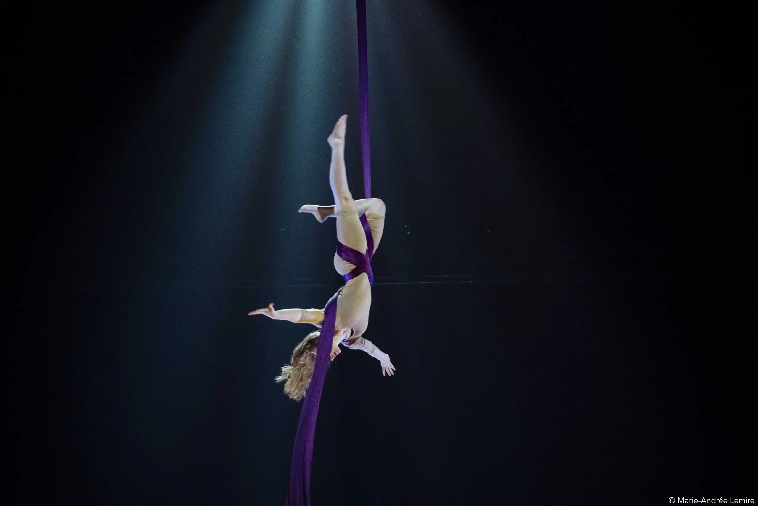 Une artiste aérienne, Margaux Wasteels, est suspendue à l'envers dans les airs, enveloppée dans un tissu de soie violet, sous les projecteurs qui l'éclairent sur une scène sombre.