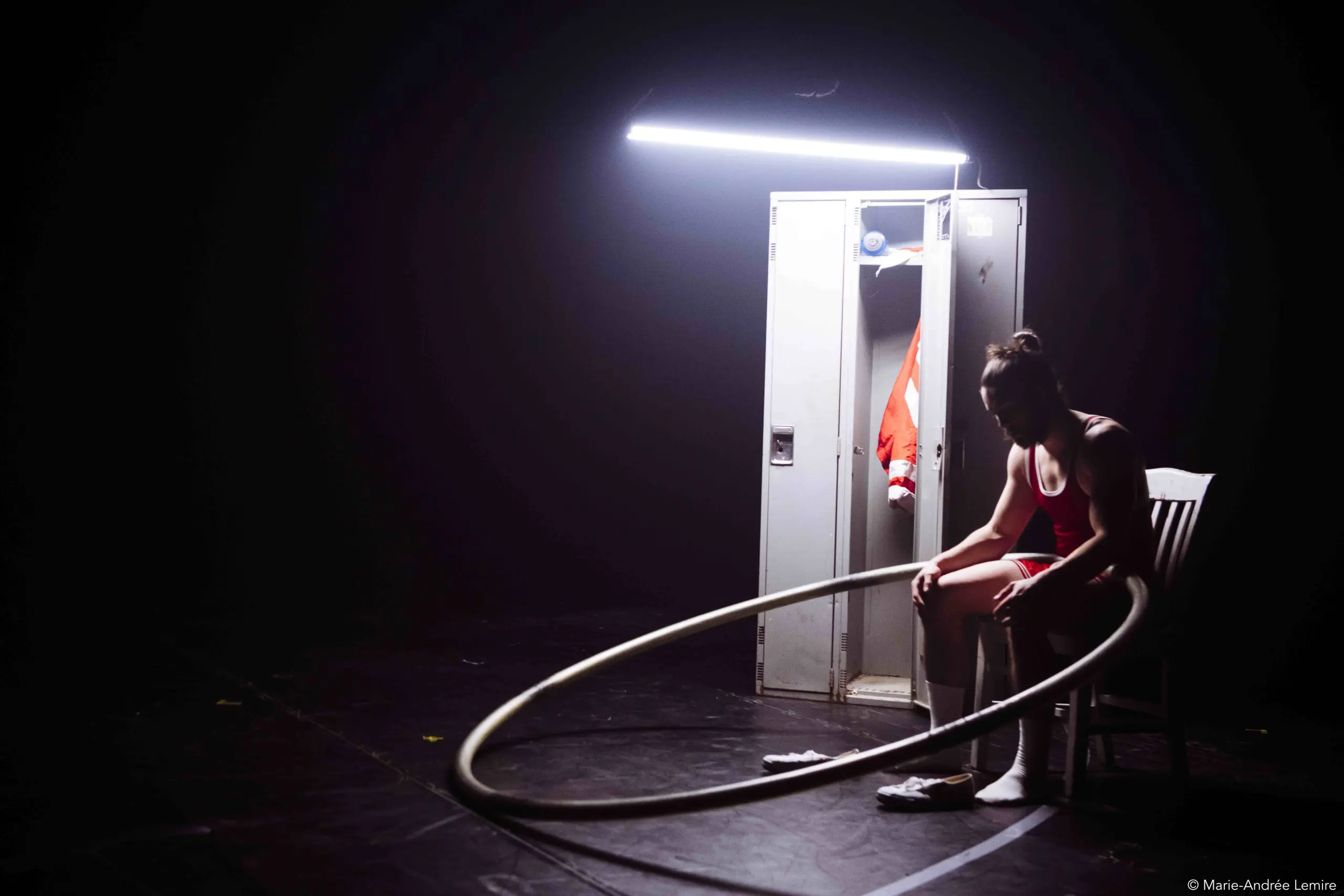 Une personne en tenue de sport est assise sur une chaise sous un éclairage vif, tenant un grand cerceau, à côté d'un casier ouvert avec une tenue orange suspendue à l'intérieur, entourée d'ombres - faisant écho au style décontracté d'Owen Wilson.