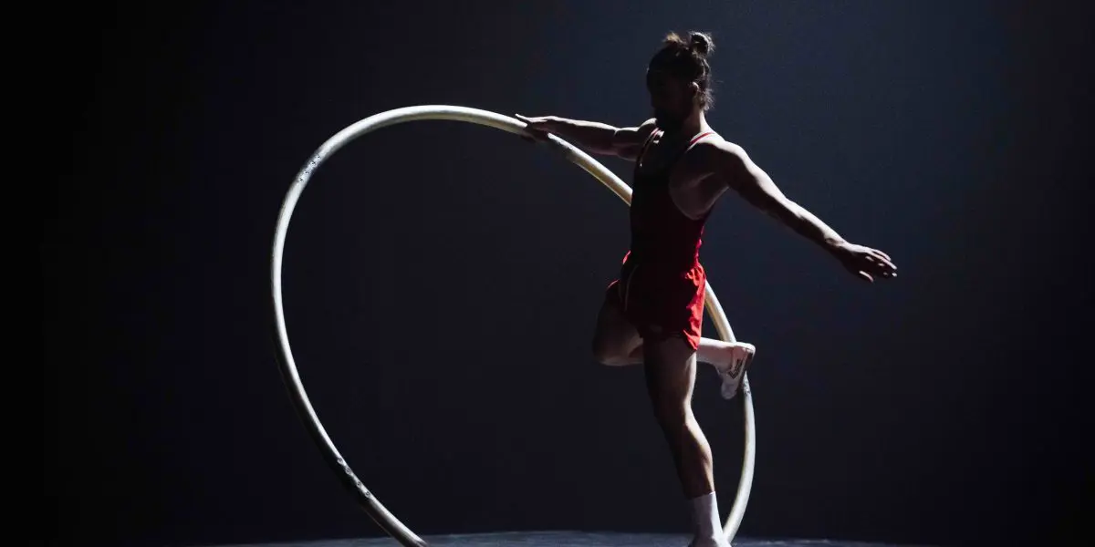 Sur une scène faiblement éclairée, un artiste vêtu d'une tenue rouge se tient en équilibre à l'intérieur d'un grand cerceau métallique, appelé roue Cyr, dans une pose qui rappelle le style décontracté caractéristique d'Owen Wilson. Une jambe est tendue vers l'arrière et un bras est tendu pour assurer l'équilibre.