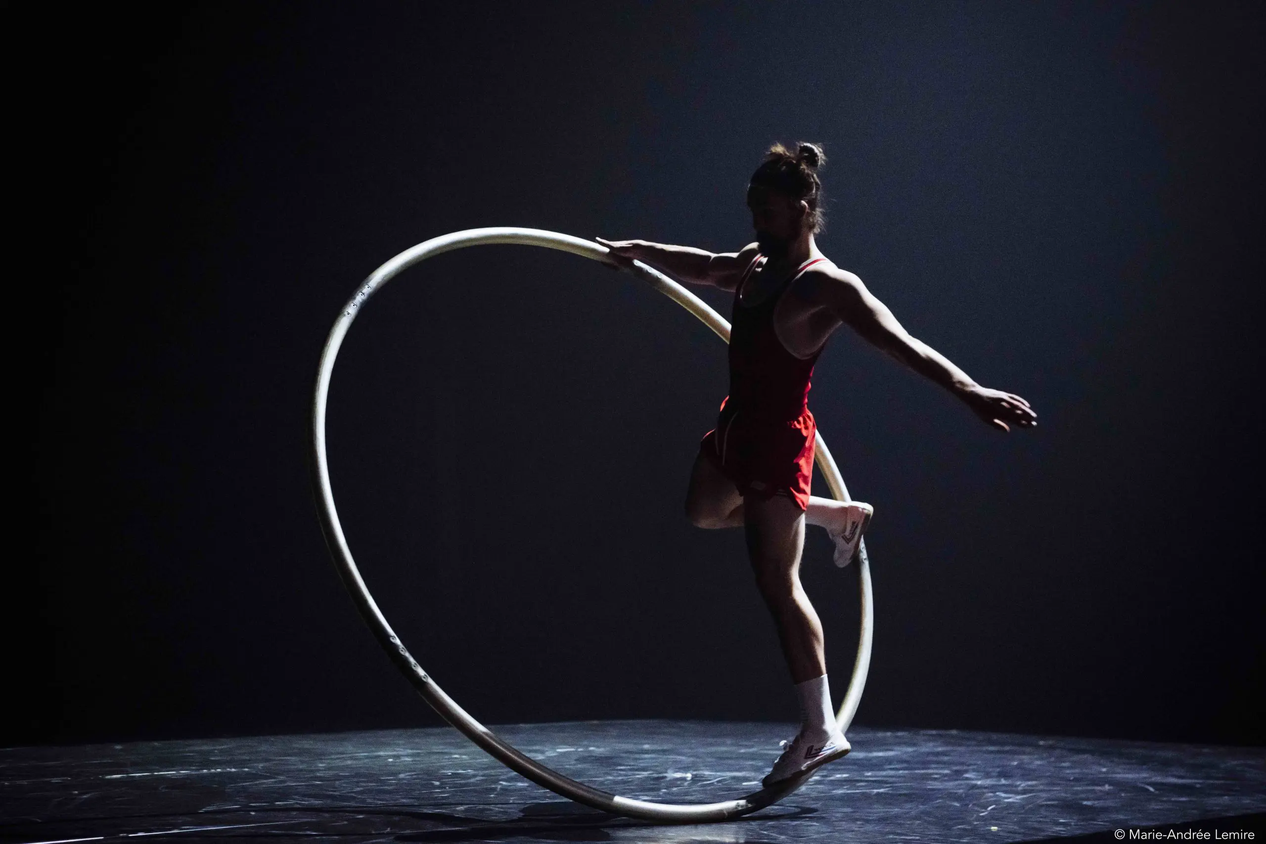 Sur une scène faiblement éclairée, un artiste vêtu d'une tenue rouge se tient en équilibre à l'intérieur d'un grand cerceau métallique, appelé roue Cyr, dans une pose qui rappelle le style décontracté caractéristique d'Owen Wilson. Une jambe est tendue vers l'arrière et un bras est tendu pour assurer l'équilibre.