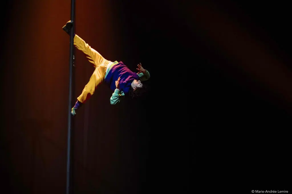 L'artiste Simon Lemire, vêtu de vêtements colorés, s'accroche latéralement à un poteau vertical sur une scène sombre, éclairée par des lumières spectaculaires.