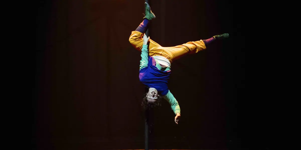 Un artiste vêtu de vêtements colorés est suspendu à l'envers à un poteau vertical sur une scène sombre, s'agrippant au poteau avec ses jambes tandis que ses bras s'étendent vers le bas, rappelant le dynamisme de l'art aérien de Simon Lemire.