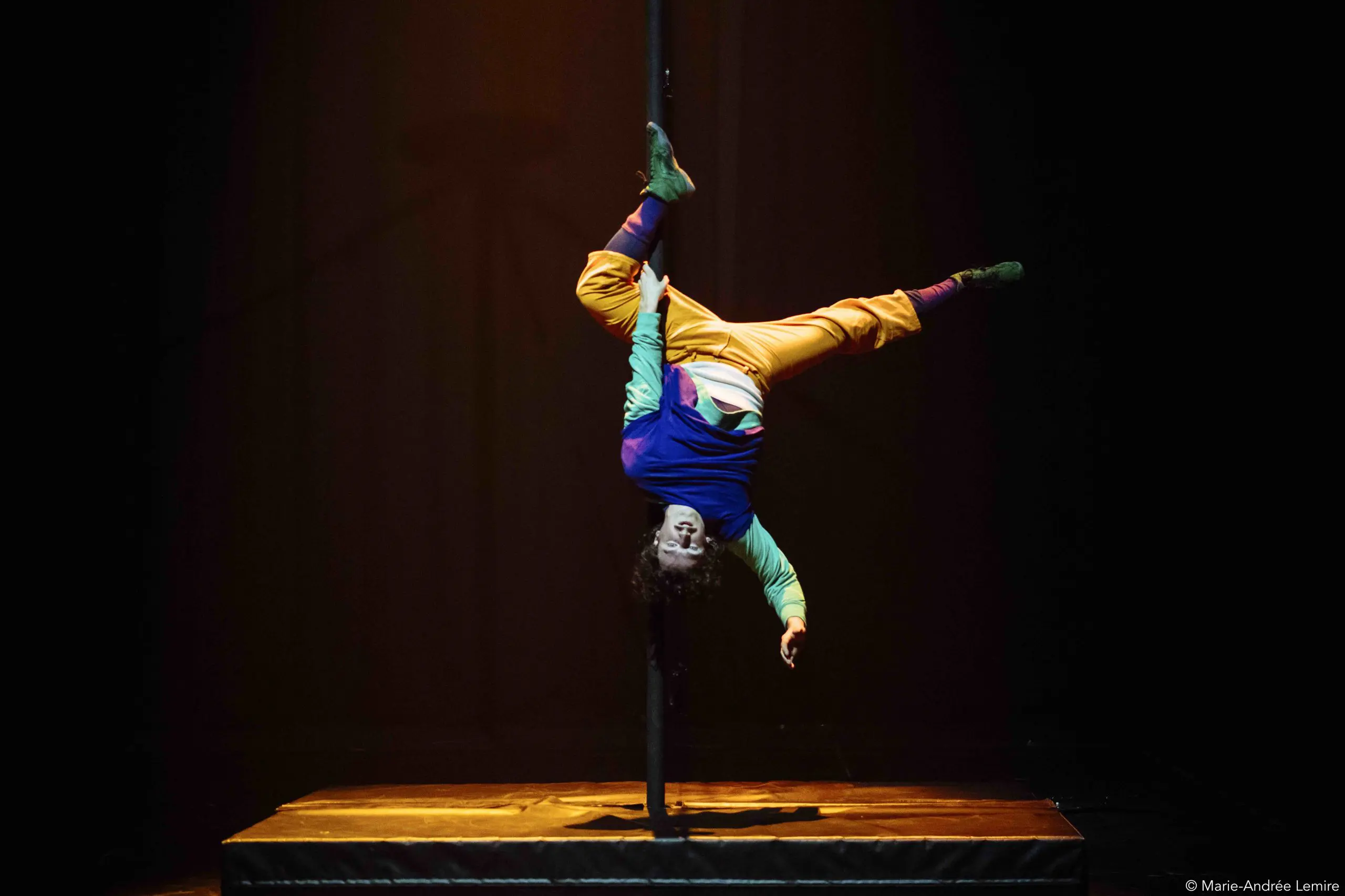 Un artiste vêtu de vêtements colorés est suspendu à l'envers à un poteau vertical sur une scène sombre, s'agrippant au poteau avec ses jambes tandis que ses bras s'étendent vers le bas, rappelant le dynamisme de l'art aérien de Simon Lemire.