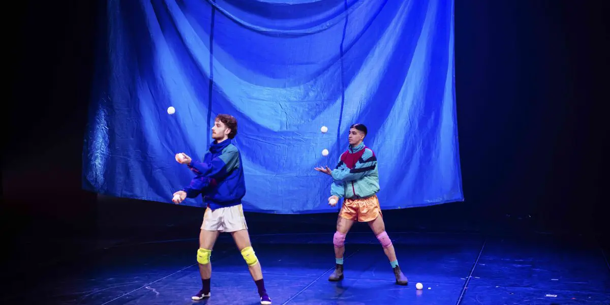 Deux artistes, Zachary Martel et Santiago Rivera Laugerud, vêtus de tenues de sport colorées et de genouillères, jonglent avec des balles blanches sur une scène faiblement éclairée, avec une grande toile de fond bleue suspendue derrière eux.