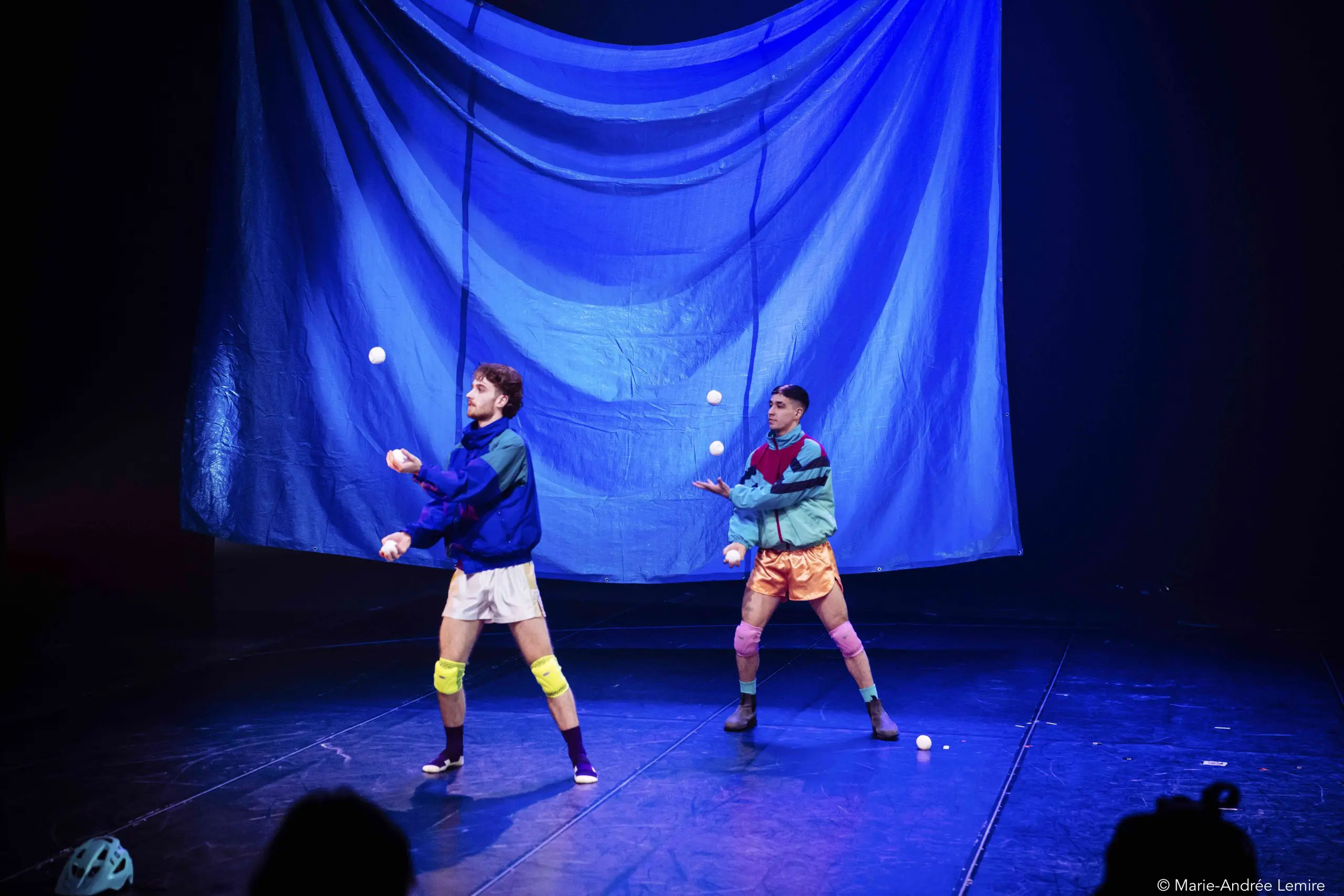 Deux artistes, Zachary Martel et Santiago Rivera Laugerud, vêtus de tenues de sport colorées et de genouillères, jonglent avec des balles blanches sur une scène faiblement éclairée, avec une grande toile de fond bleue suspendue derrière eux.