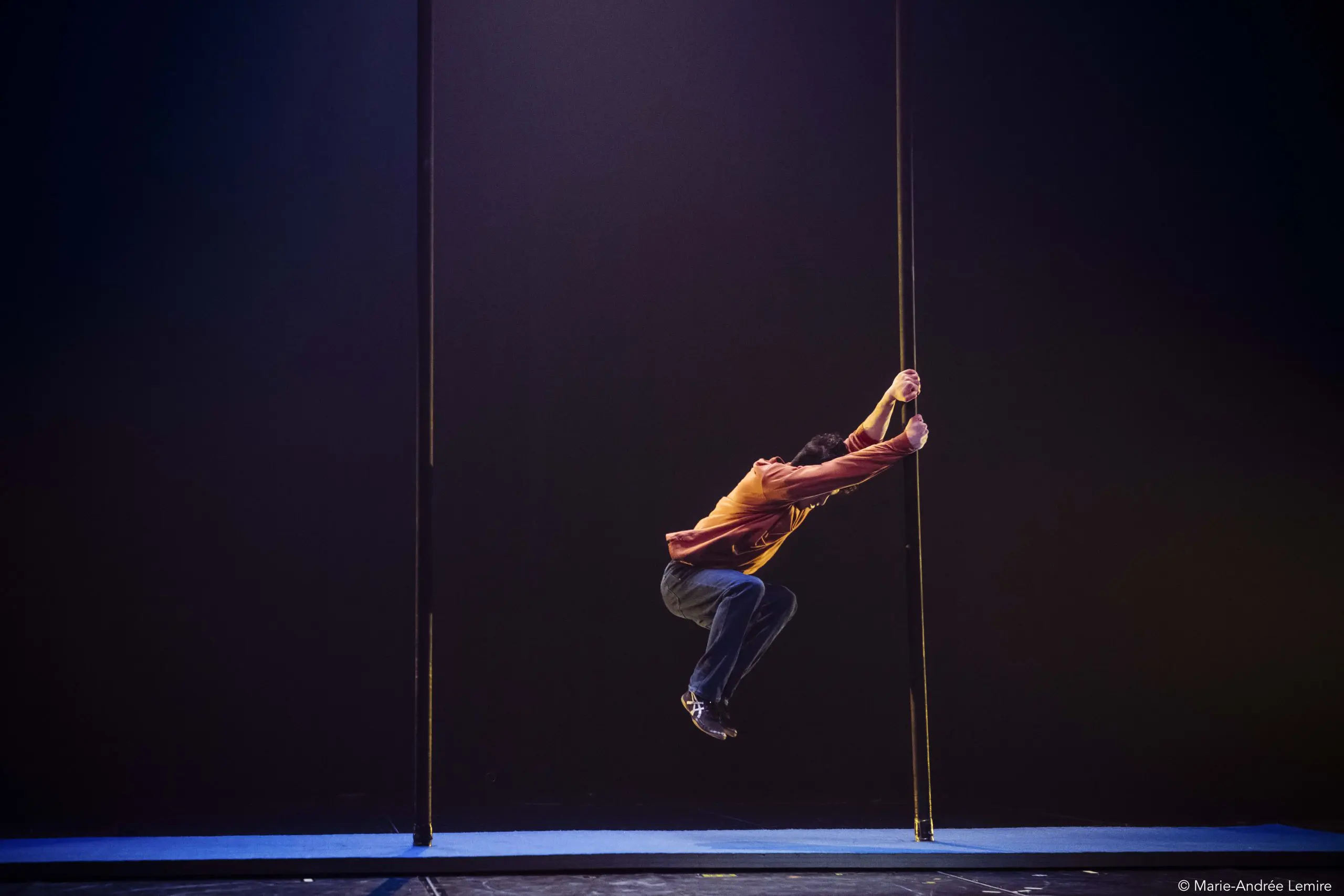 Une personne vêtue d'un pull rouge et d'un pantalon sombre exécute un mouvement acrobatique, saisissant deux poteaux verticaux et suspendant son corps au-dessus d'une scène à l'éclairage dramatique, rappelant le style caractéristique de Teo Le Baut.