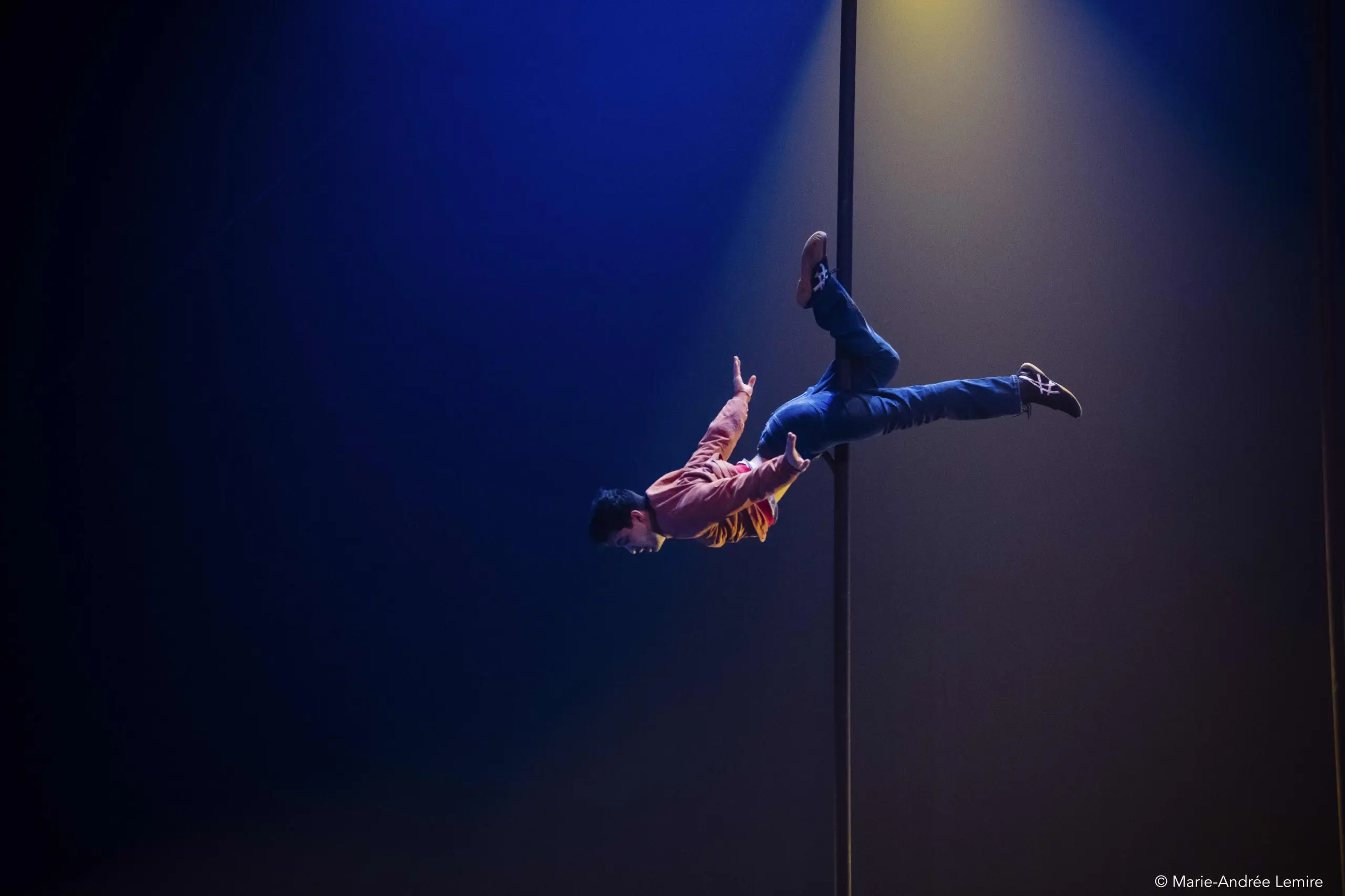 L'artiste Teo Le Baut, vêtu d'une chemise brune et d'un jean, est suspendu horizontalement à un poteau vertical, se tenant en équilibre avec un bras sous un projecteur dramatique sur un fond sombre et éclairé en bleu.