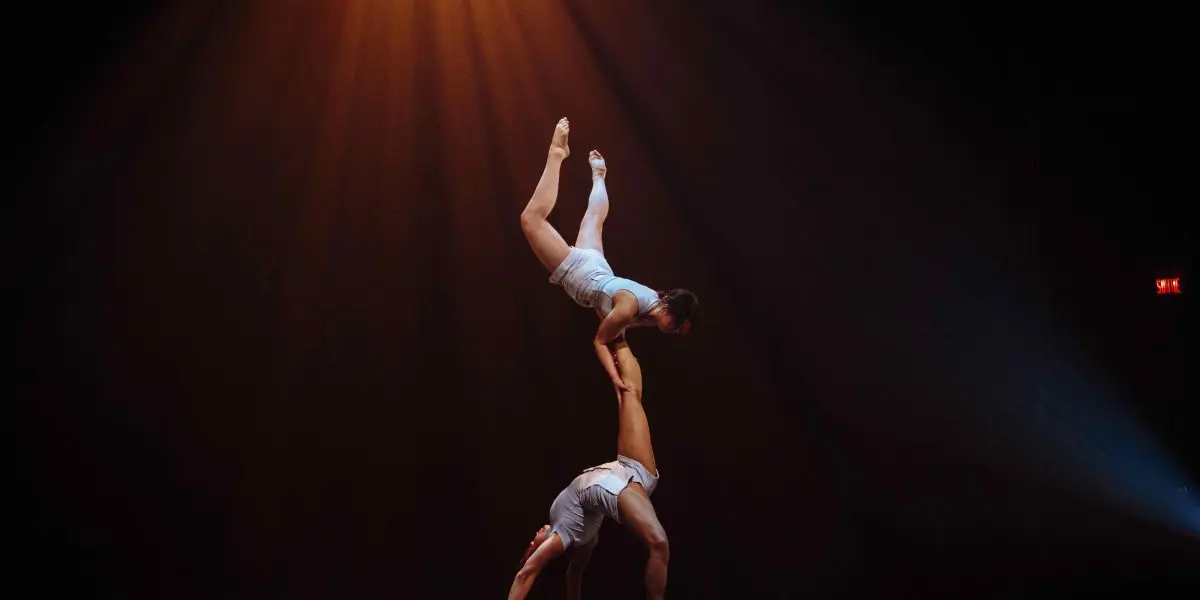 Deux acrobates, Léann Gingras et Kaisha Dessalines Wright, en tenue blanche, exécutent un numéro d'équilibre sur une scène sombre sous un projecteur lumineux, l'un debout et tenant l'autre à l'envers au-dessus de sa tête.