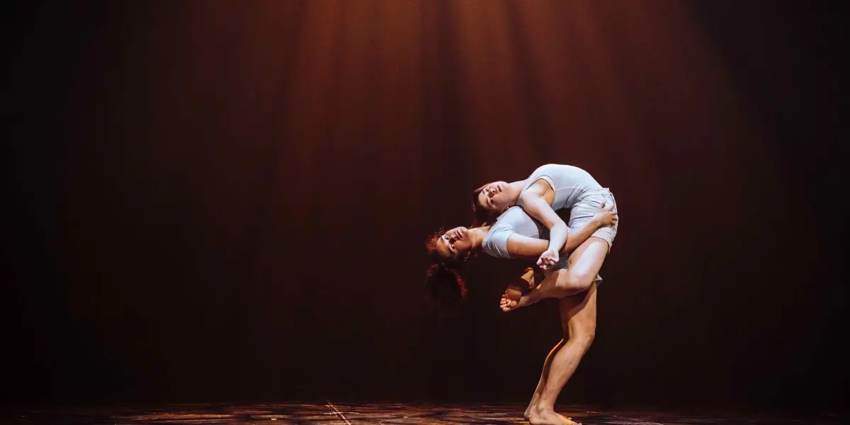 Deux danseuses contemporaines, Léann Gingras et Kaisha Dessalines Wright, en tenues blanches, exécutent un duo expressif sur une scène faiblement éclairée, l'une se soulevant et s'arquant vers l'arrière au-dessus de l'autre sous la lumière chaleureuse des projecteurs.