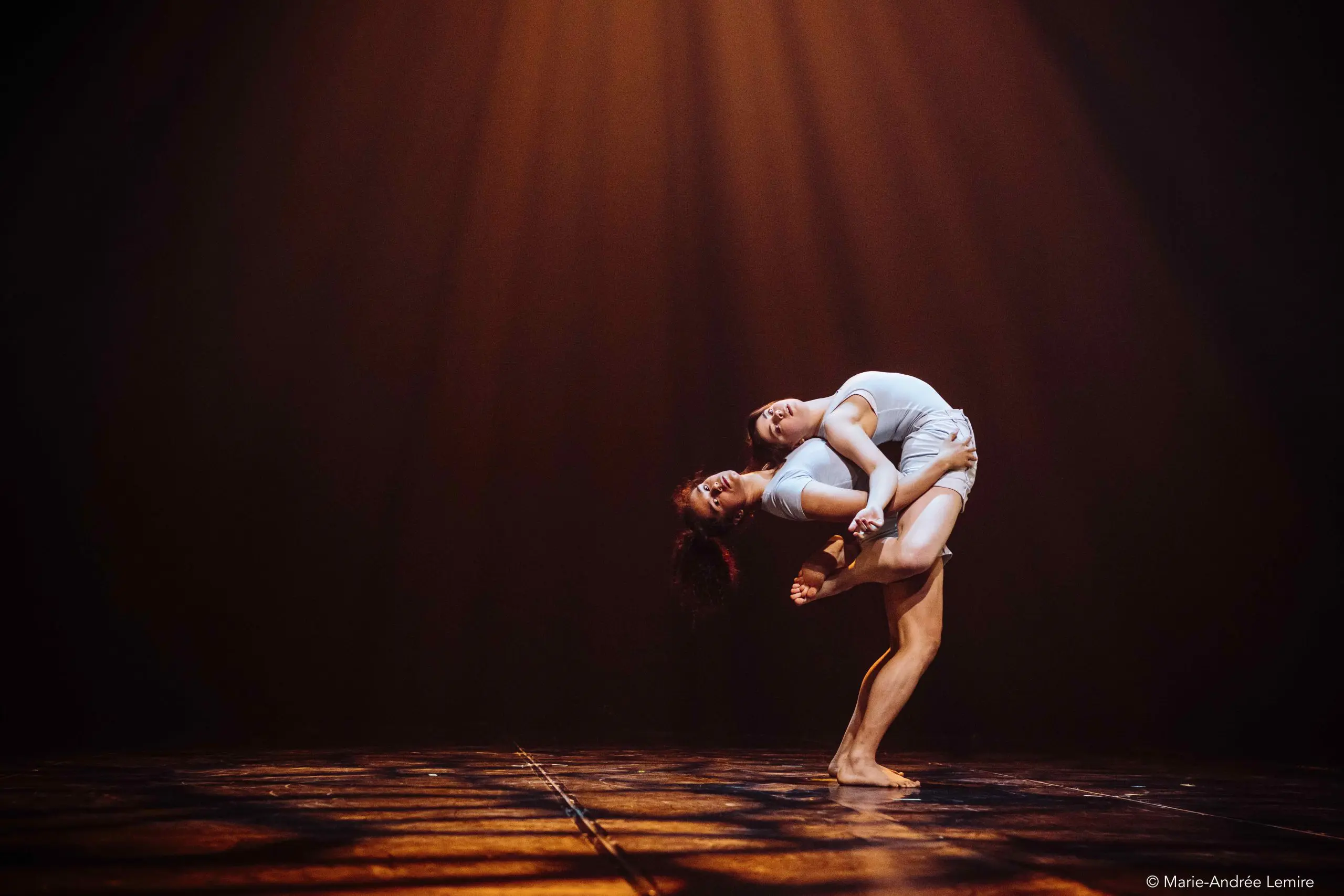 Deux danseuses contemporaines, Léann Gingras et Kaisha Dessalines Wright, en tenues blanches, exécutent un duo expressif sur une scène faiblement éclairée, l'une se soulevant et s'arquant vers l'arrière au-dessus de l'autre sous la lumière chaleureuse des projecteurs.