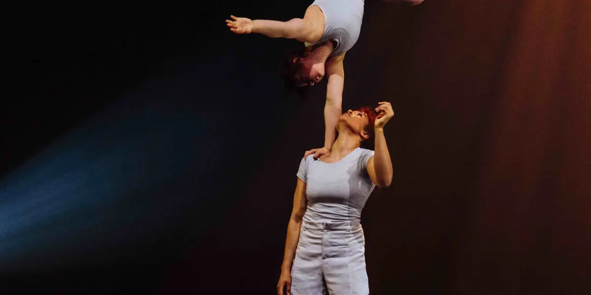 Deux acrobates, Léann Gingras et Kaisha Dessalines Wright, se produisent sur scène, l'une en équilibre sur la main de l'autre. Toutes deux portent des tenues blanches et l'éclairage dramatique de la scène met en valeur leur pose saisissante.