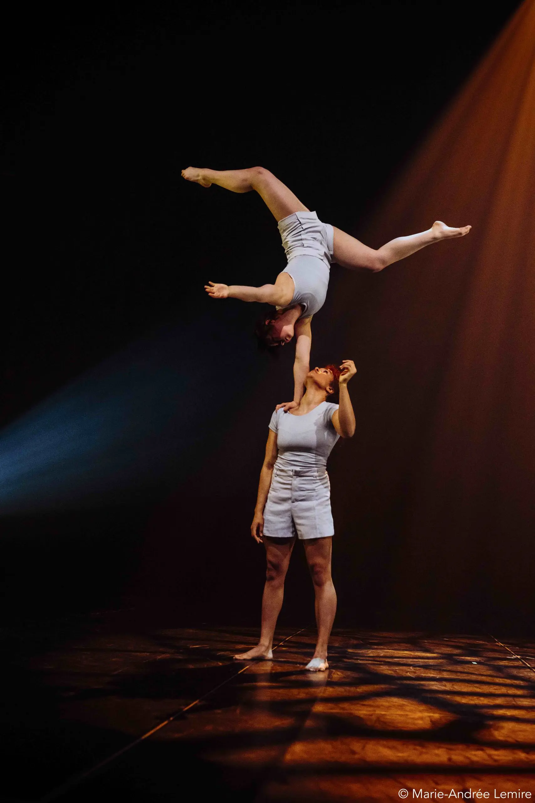 Deux acrobates, Léann Gingras et Kaisha Dessalines Wright, se produisent sur scène, l'une en équilibre sur la main de l'autre. Toutes deux portent des tenues blanches et l'éclairage dramatique de la scène met en valeur leur pose saisissante.