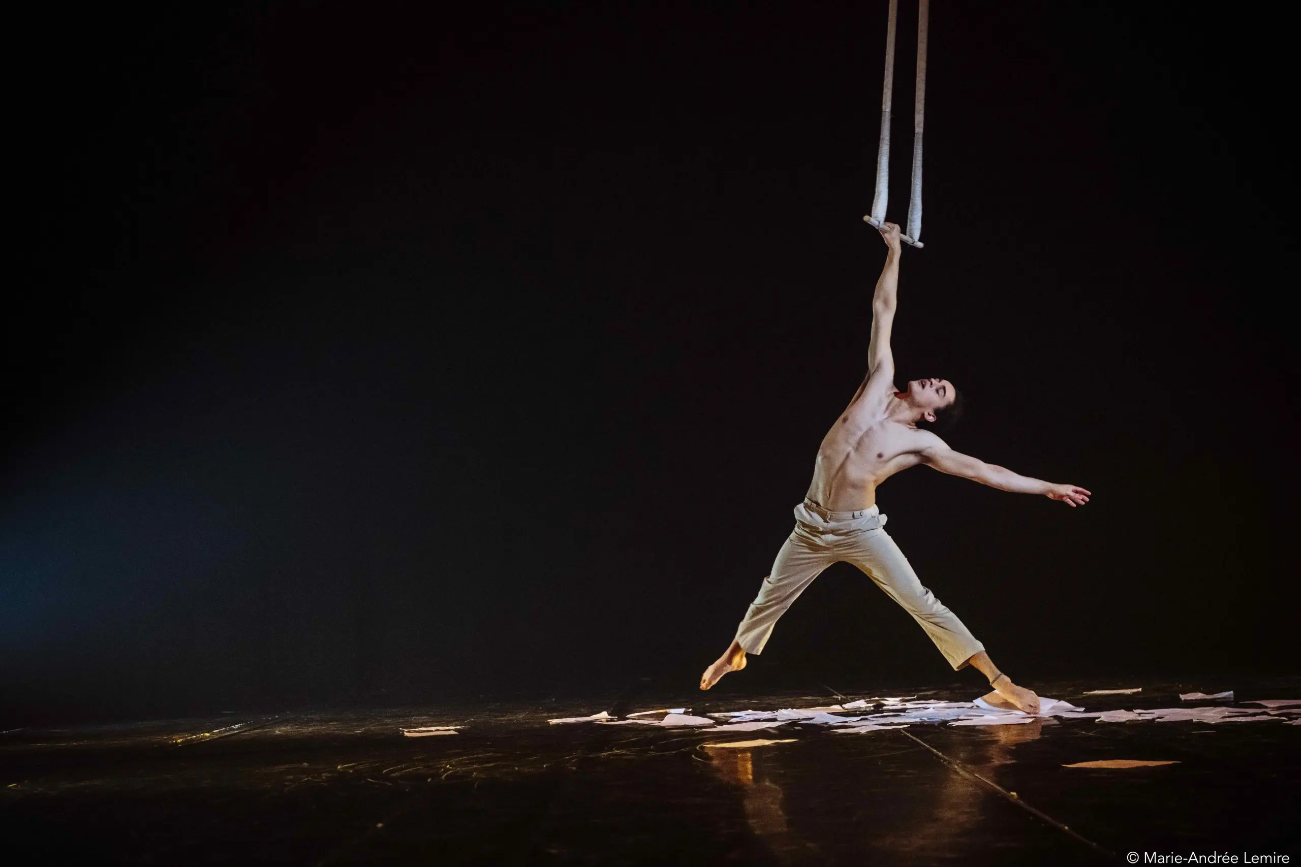 Zébulon Simoneau, torse nu et en pantalon clair, se produit sur scène, gracieusement suspendu par un bras à des sangles aériennes au-dessus de papiers épars, sur un fond sombre et dramatique.