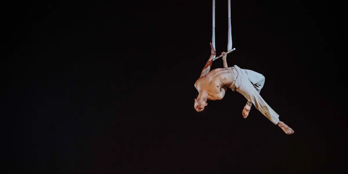 Un artiste de cirque, Zebulon Simoneau, exécute un numéro aérien sur des cordes suspendues, la tête en bas dans les airs sur un fond sombre, le corps gracieusement arqué et vêtu d'un pantalon de couleur claire.