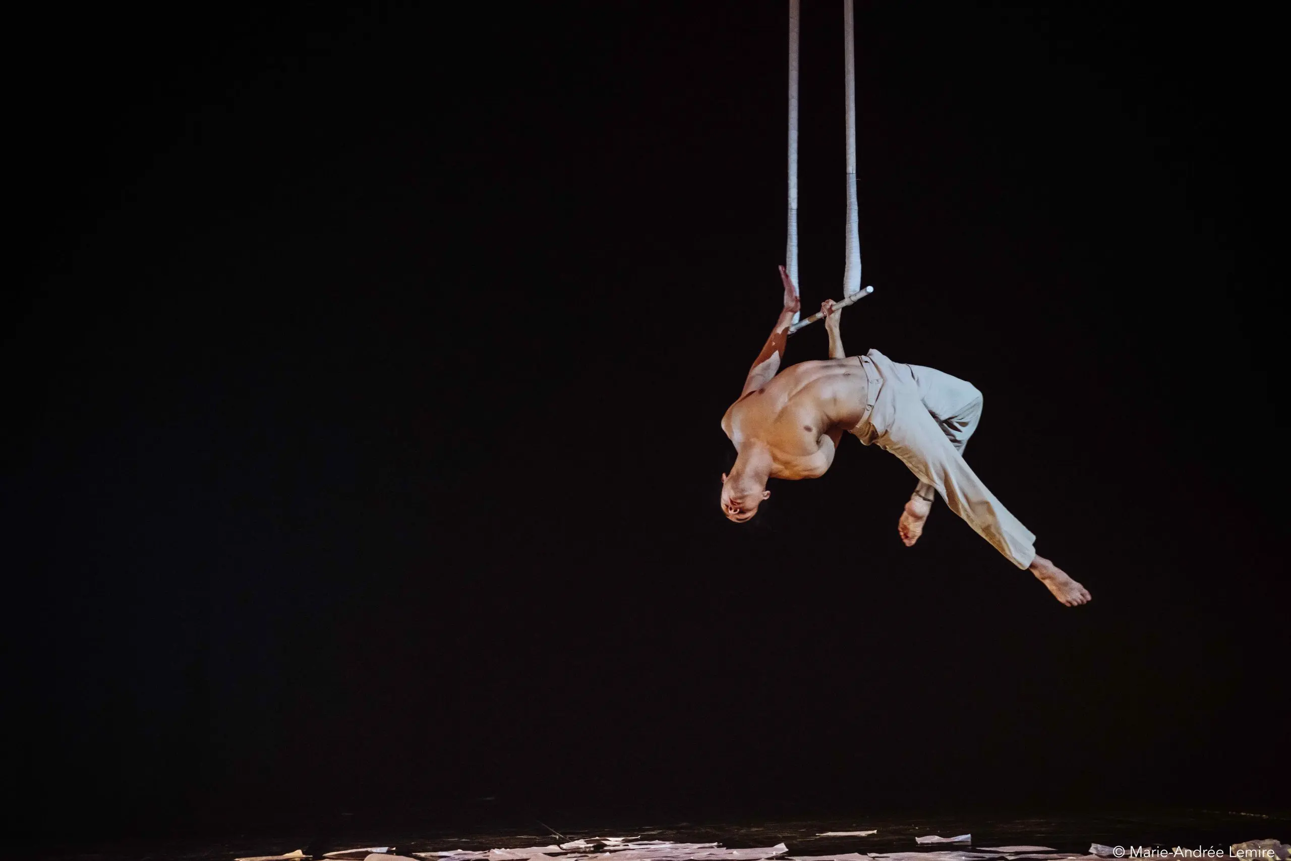 Un artiste de cirque, Zebulon Simoneau, exécute un numéro aérien sur des cordes suspendues, la tête en bas dans les airs sur un fond sombre, le corps gracieusement arqué et vêtu d'un pantalon de couleur claire.