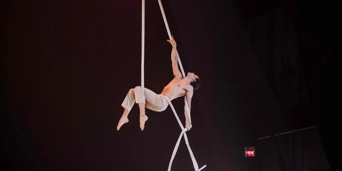 L'artiste de cirque Zebulon Simoneau, vêtu d'un pantalon blanc, se tient en équilibre sur un appareil aérien, tenant des cordes suspendues d'une main et d'un pied tout en arquant gracieusement son corps sous un éclairage de scène spectaculaire.