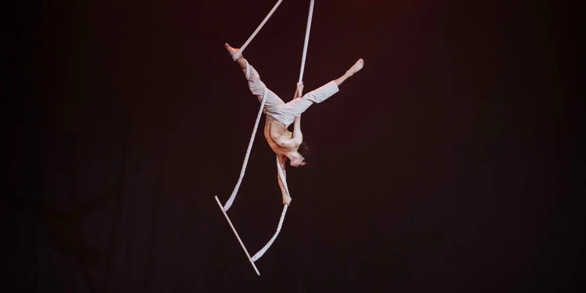 L'artiste aérien Zebulon Simoneau, en pantalon blanc, exécute un saut acrobatique sur des sangles suspendues sur un fond sombre, en équilibre à l'envers au-dessus de la scène.