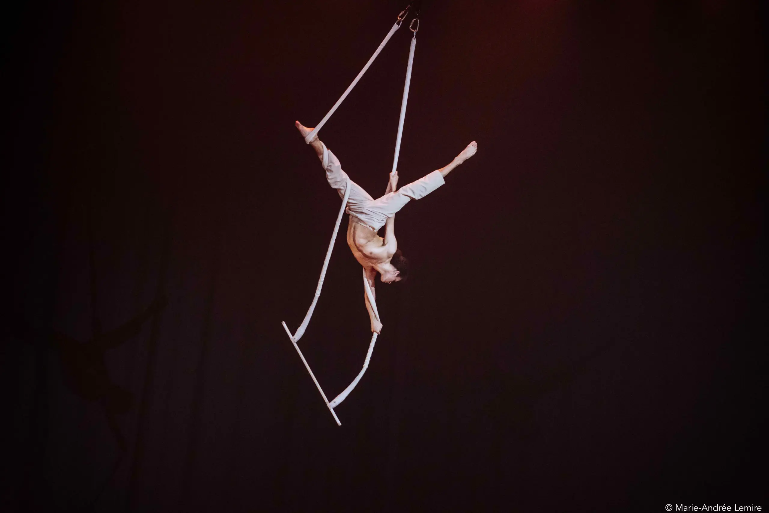 L'artiste aérien Zebulon Simoneau, en pantalon blanc, exécute un saut acrobatique sur des sangles suspendues sur un fond sombre, en équilibre à l'envers au-dessus de la scène.
