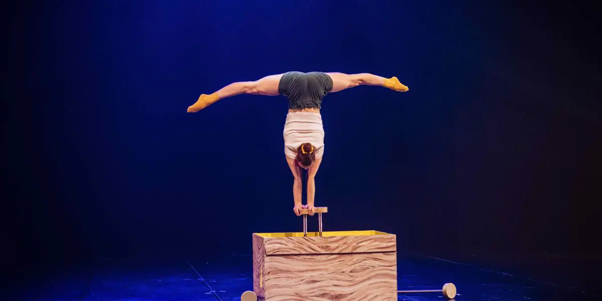 Emma Verhaeghe se tient en équilibre sur les mains au-dessus d'une caisse en bois, les jambes étendues en fente au-dessus de sa tête, sur une scène sombre éclairée d'une lumière bleue dramatique.
