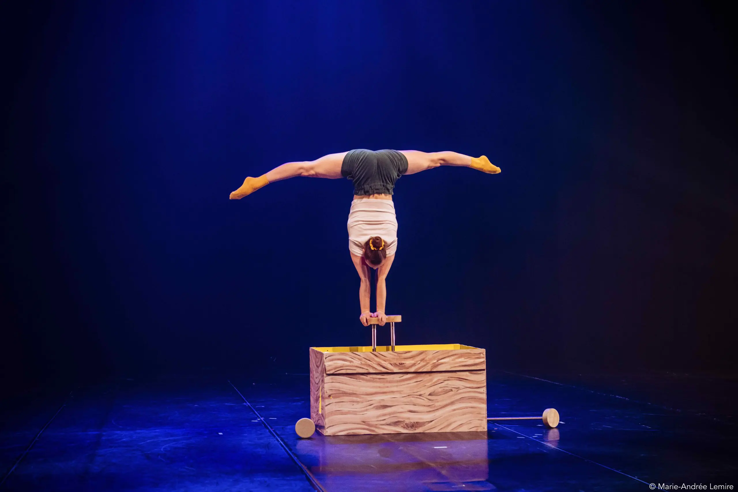 Emma Verhaeghe se tient en équilibre sur les mains au-dessus d'une caisse en bois, les jambes étendues en fente au-dessus de sa tête, sur une scène sombre éclairée d'une lumière bleue dramatique.