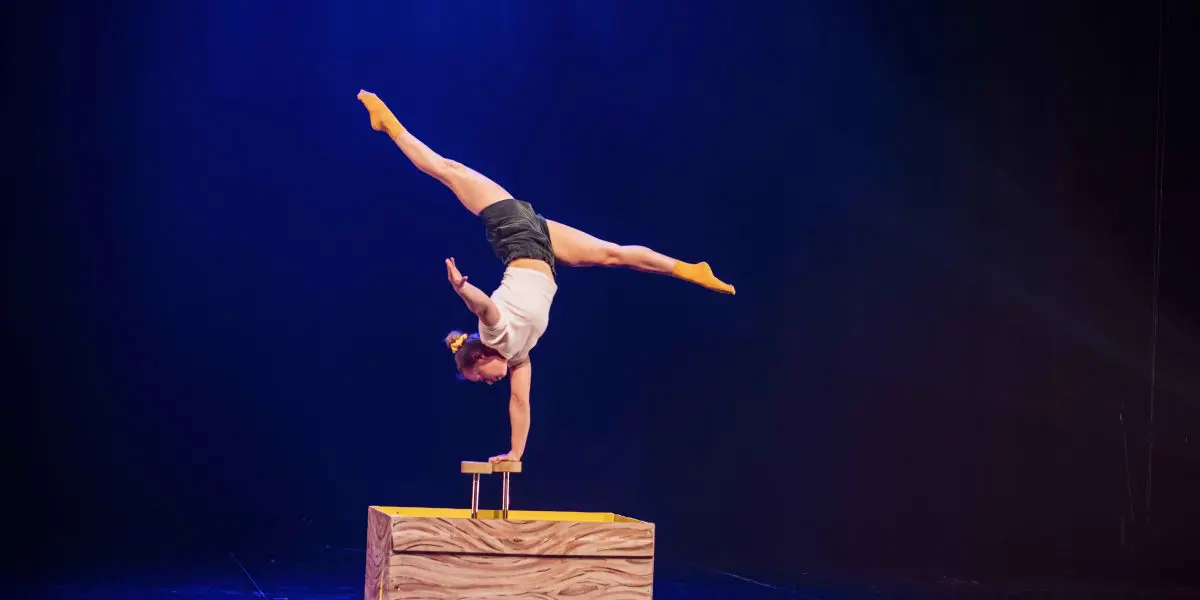 Emma Verhaeghe, une artiste de cirque, se tient en équilibre sur une main au sommet d'une plate-forme en bois, les jambes étendues en fente, avec des chaussettes jaunes et des lumières de scène soulignant l'arrière-plan sombre.