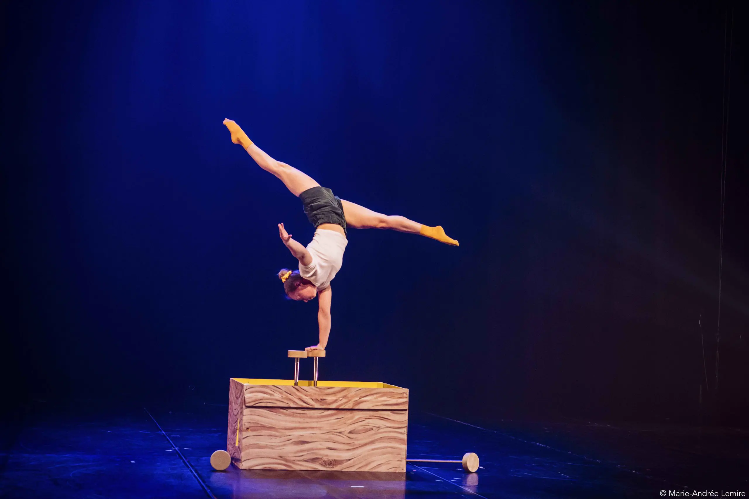 Emma Verhaeghe, une artiste de cirque, se tient en équilibre sur une main au sommet d'une plate-forme en bois, les jambes étendues en fente, avec des chaussettes jaunes et des lumières de scène soulignant l'arrière-plan sombre.