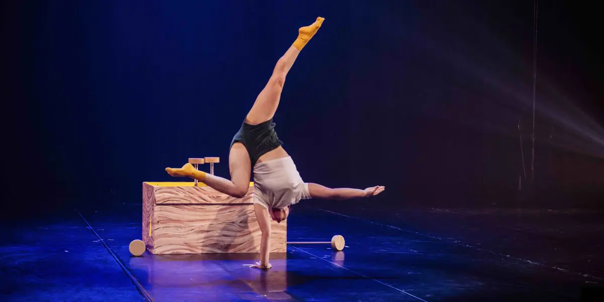 Une artiste, peut-être Emma Verhaeghe, en chaussettes jaunes et short foncé, se tient en équilibre sur une main, les jambes tendues en fente, sur une scène faiblement éclairée, avec une boîte en bois et de petits accessoires en arrière-plan.