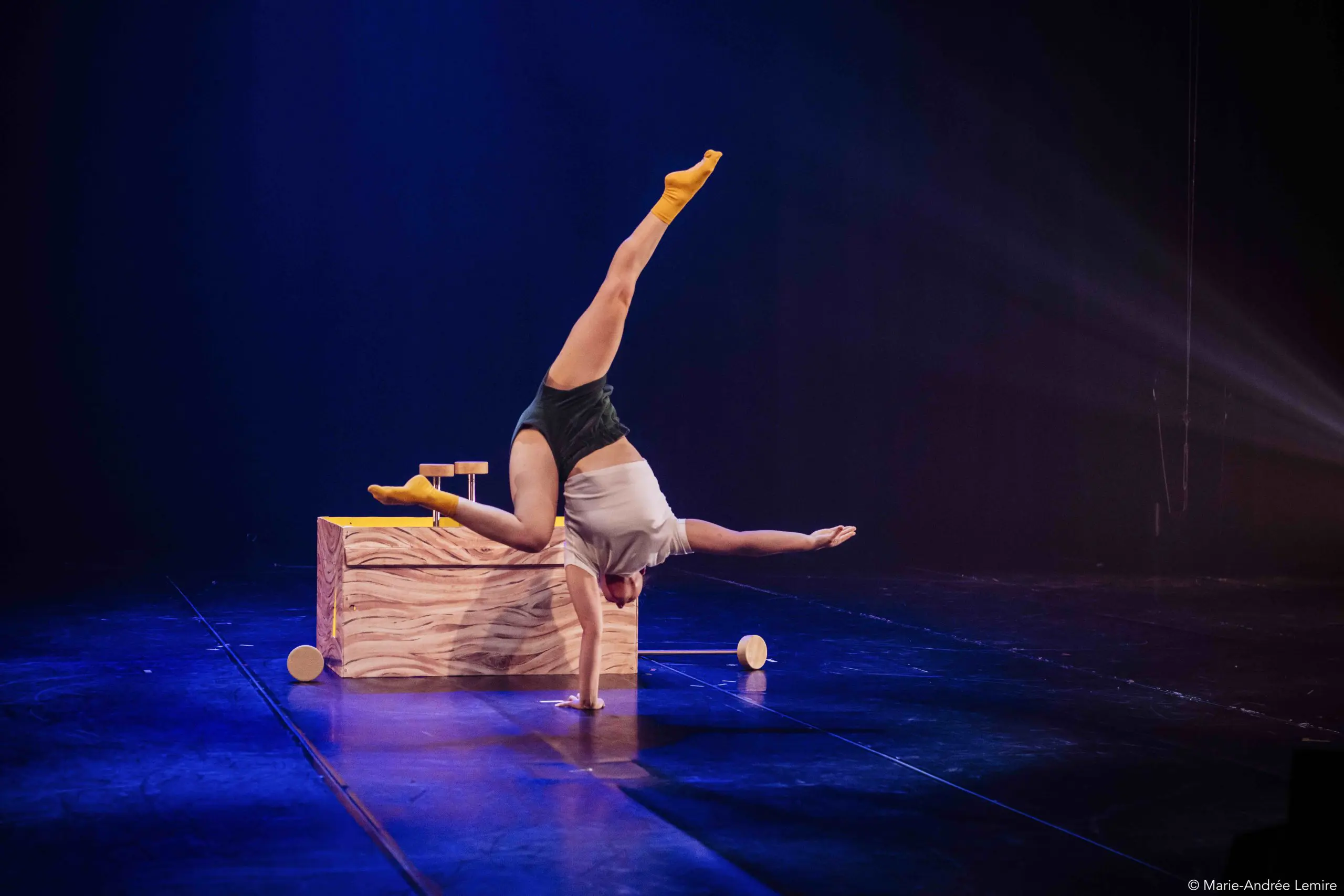 Une artiste, peut-être Emma Verhaeghe, en chaussettes jaunes et short foncé, se tient en équilibre sur une main, les jambes tendues en fente, sur une scène faiblement éclairée, avec une boîte en bois et de petits accessoires en arrière-plan.