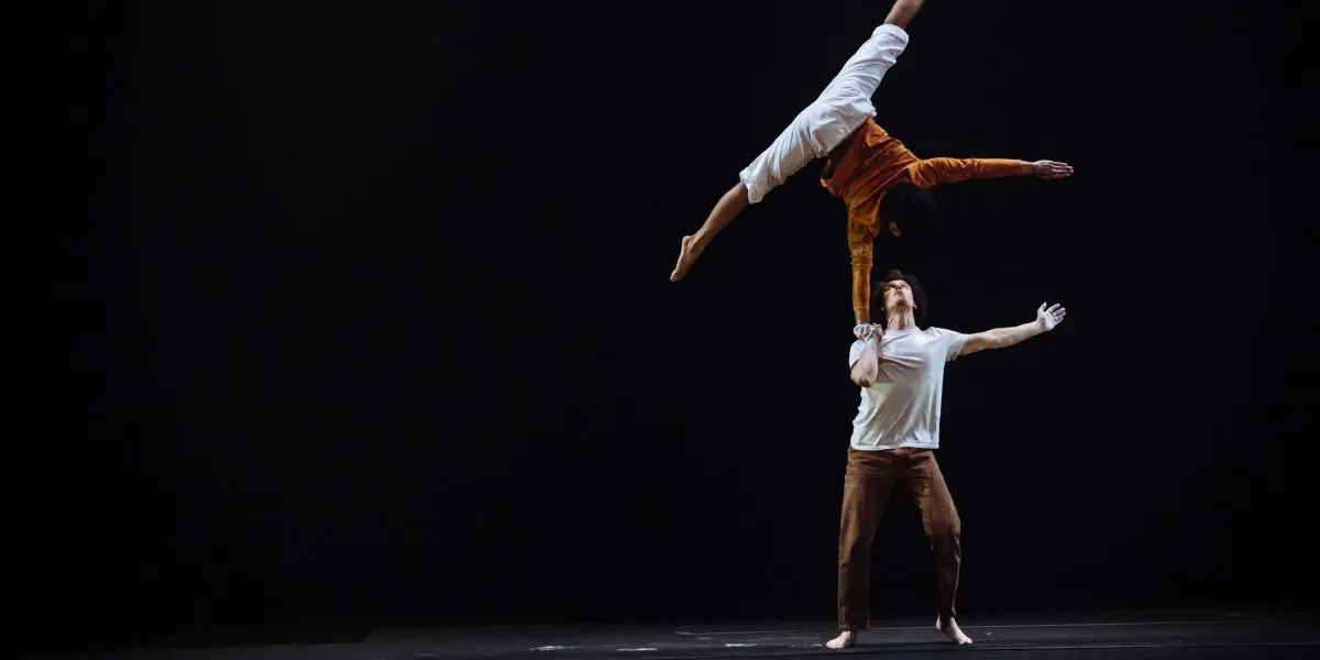 Deux acrobates se produisent sur une scène sombre ; Oliver Layher se tient debout et soulève son partenaire en l'air d'une main, tandis que la personne soulevée étend ses bras et ses jambes à l'horizontale dans une pose dramatique.