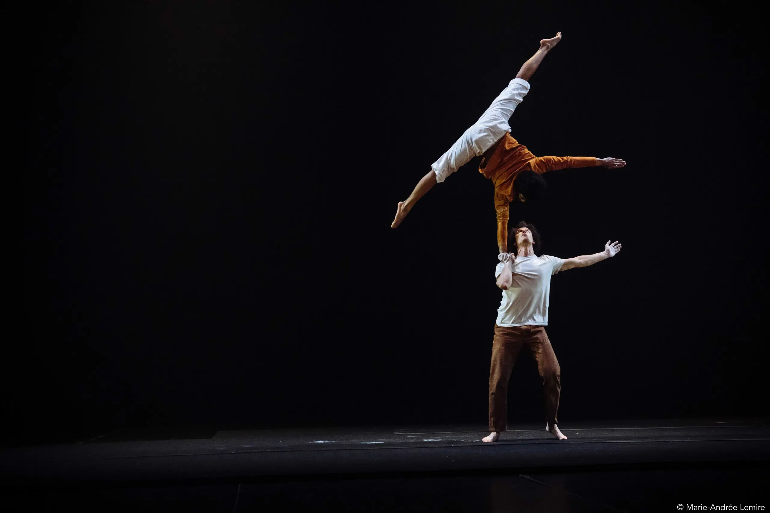Deux acrobates se produisent sur une scène sombre ; Oliver Layher se tient debout et soulève son partenaire en l'air d'une main, tandis que la personne soulevée étend ses bras et ses jambes à l'horizontale dans une pose dramatique.