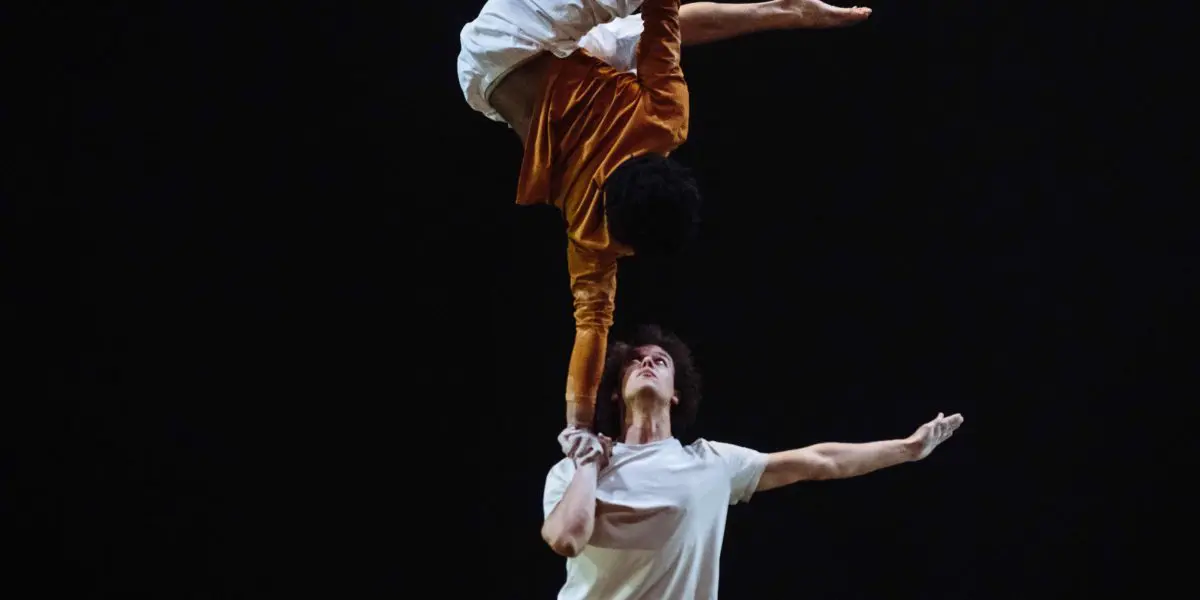 Deux artistes sur scène : Oliver Layher se tient debout, les bras tendus, et tient l'autre en équilibre sur une main, la tête en bas. L'artiste en équilibre porte un pantalon blanc et un haut orange, contrastant avec le fond sombre.