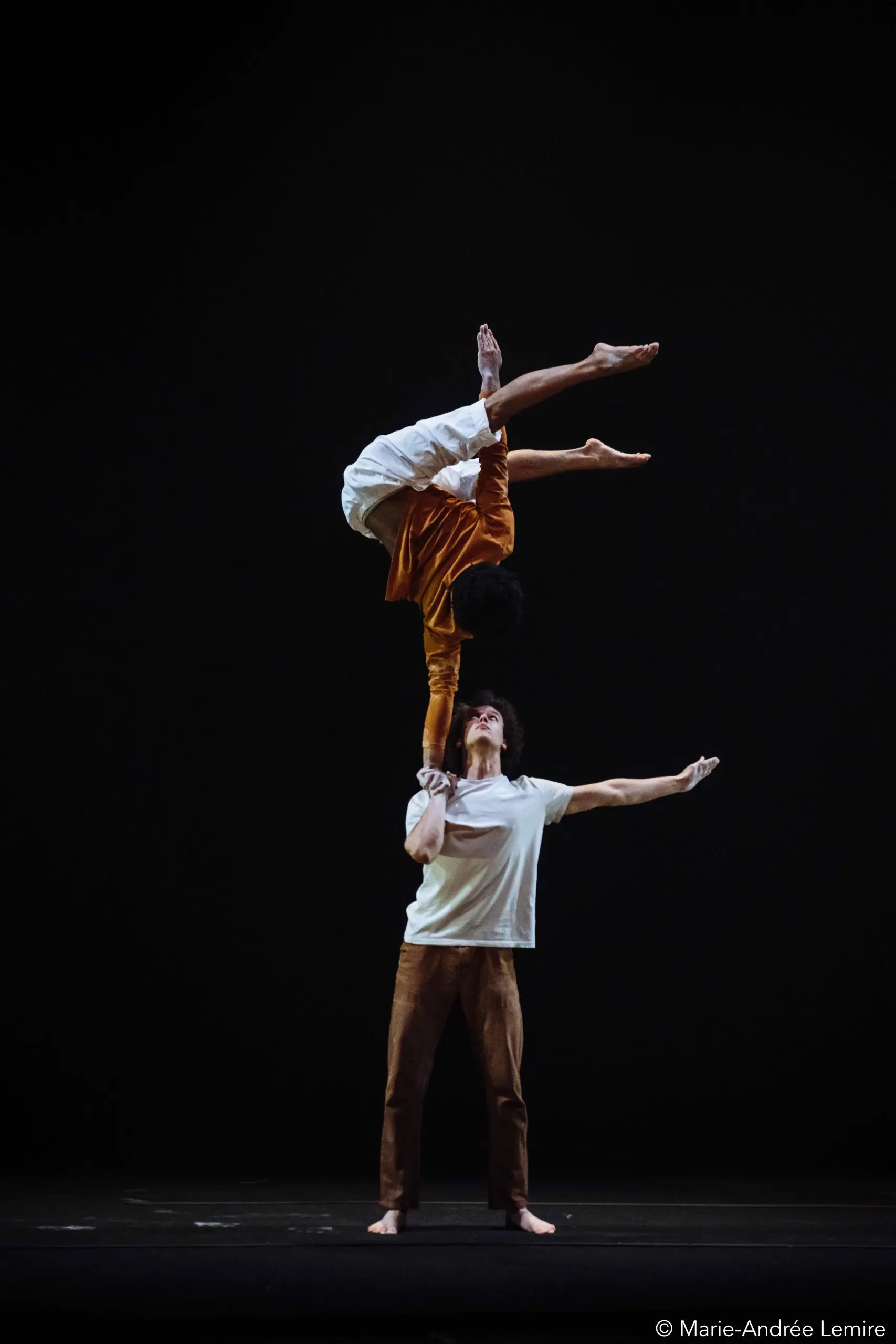 Deux acrobates, dont Kyran Lee Walton, se produisent sur scène ; l'un est en équilibre sur le bras tendu de l'autre. La base est haute et les jambes du voltigeur s'étendent gracieusement dans les airs sur un fond sombre et minimal.