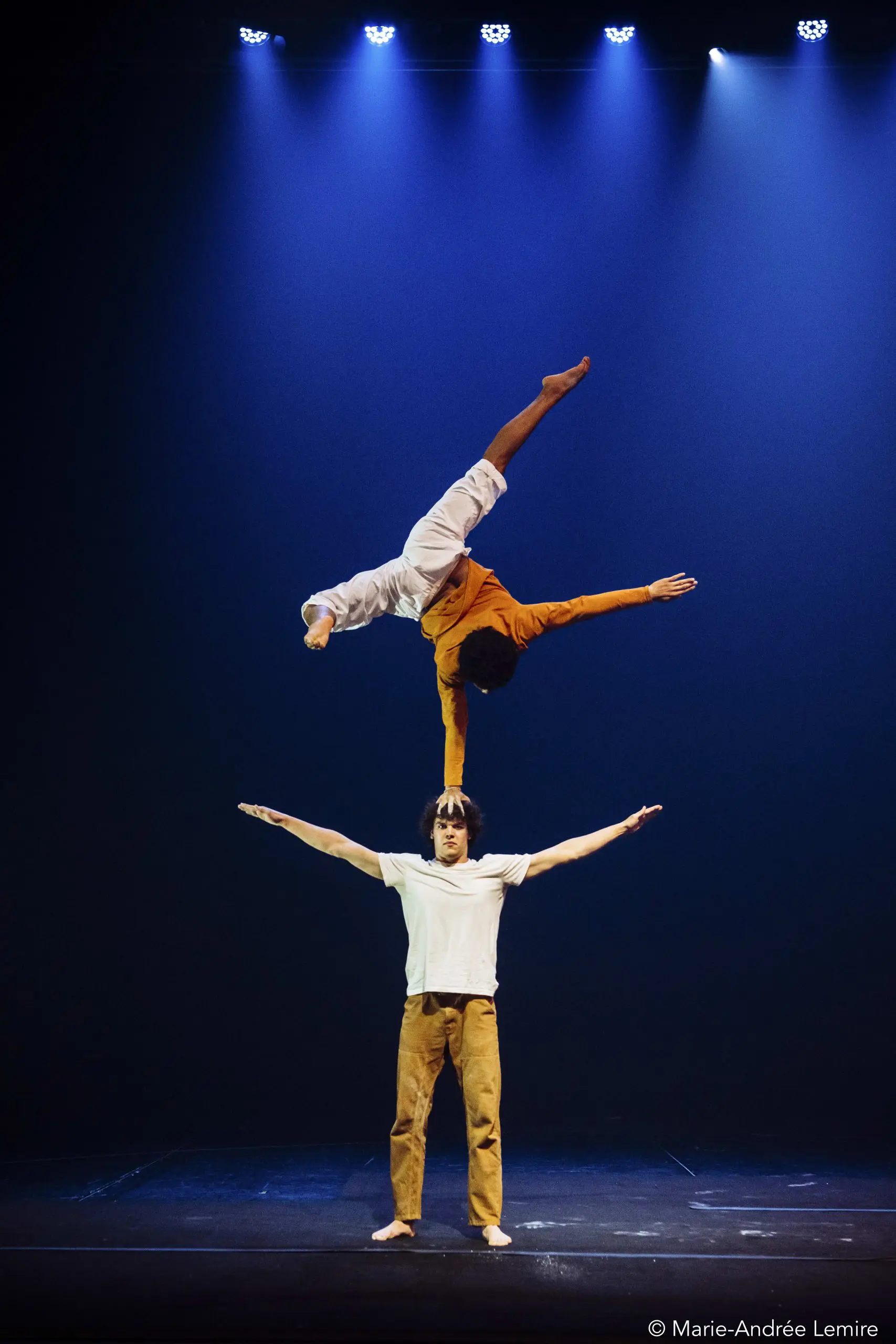 Oliver Layher se tient debout, les bras tendus, et fait tenir une acrobate à l'envers sur une main au-dessus de sa tête, sous un éclairage dramatique.