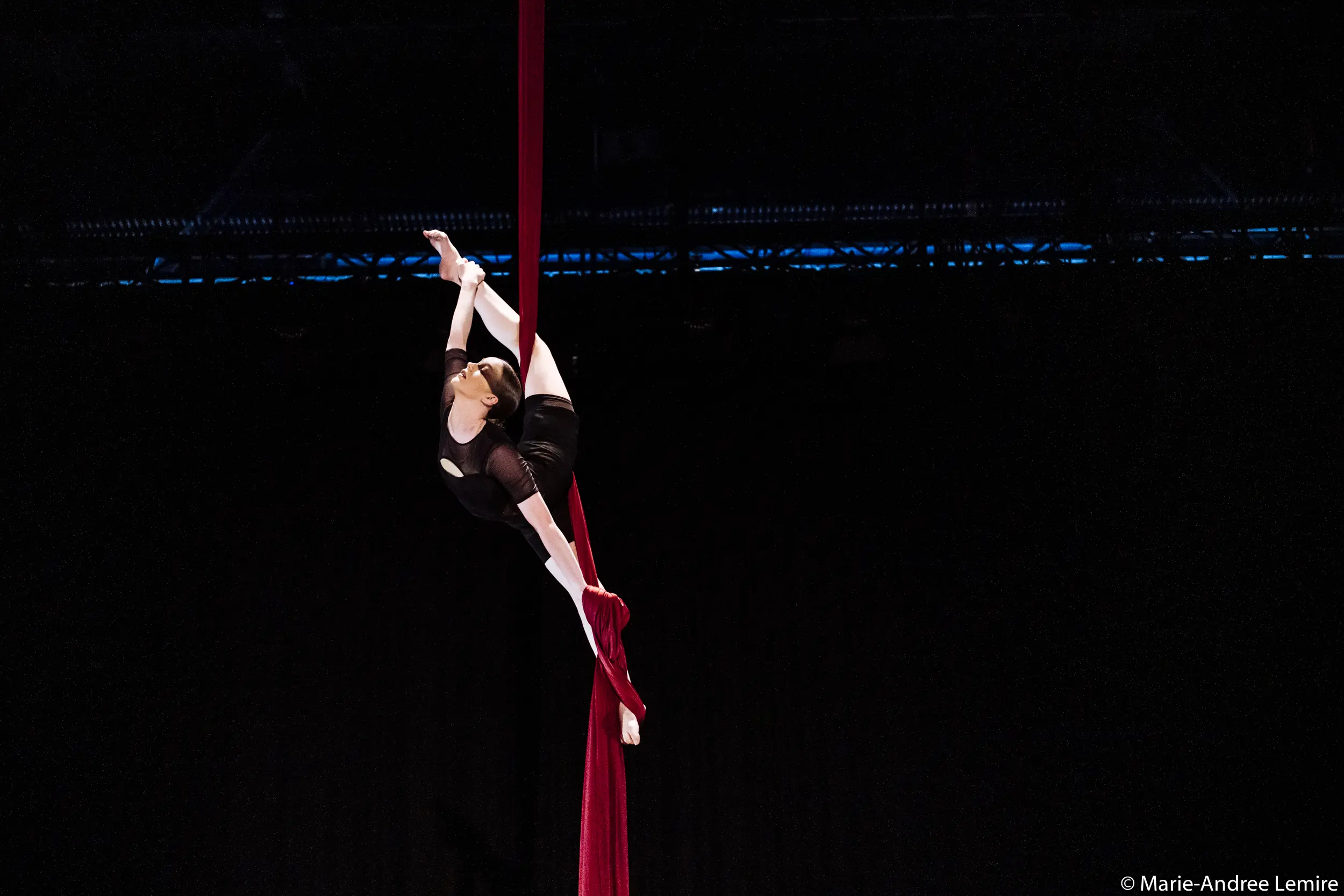 Jasmine Gagné-Morin, en costume noir, est suspendue au-dessus de la scène, étirant gracieusement ses jambes et ses bras tout en s'accrochant à des soies rouges sur un fond sombre.