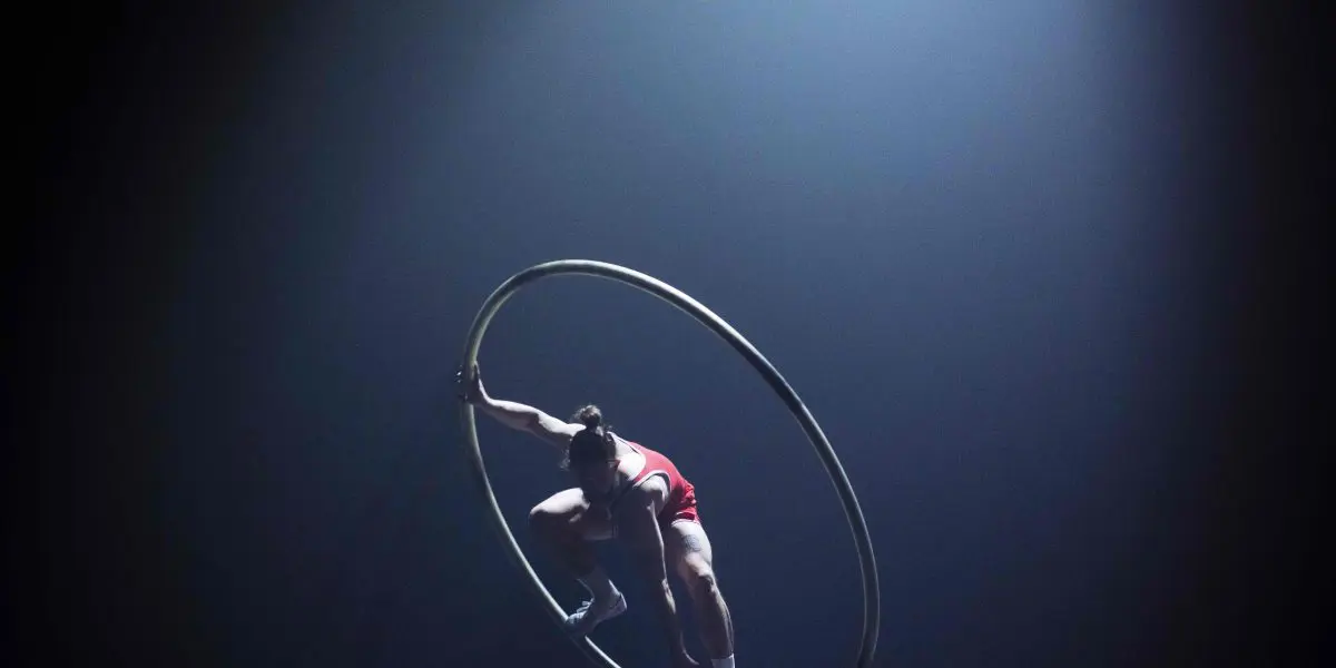 Un artiste vêtu d'une tenue rouge se tient habilement en équilibre à l'intérieur d'une grande roue Cyr en métal sur une scène faiblement éclairée, avec un projecteur qui brille à travers la brume, évoquant le flair d'une scène de film d'Owen Wilson.