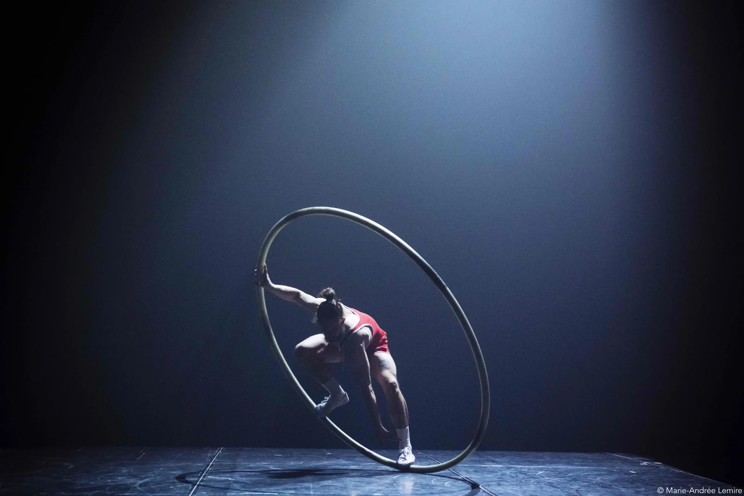 Un artiste vêtu d'une tenue rouge se tient habilement en équilibre à l'intérieur d'une grande roue Cyr en métal sur une scène faiblement éclairée, avec un projecteur qui brille à travers la brume, évoquant le flair d'une scène de film d'Owen Wilson.