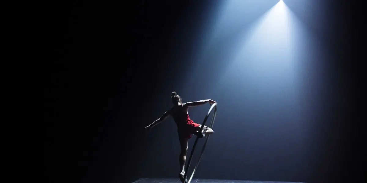 Un artiste se tient gracieusement en équilibre sur une roue Cyr sous un projecteur dramatique sur une scène sombre, sa silhouette évoquant le charisme d'Owen Wilson sur l'arrière-plan aux tons bleus.