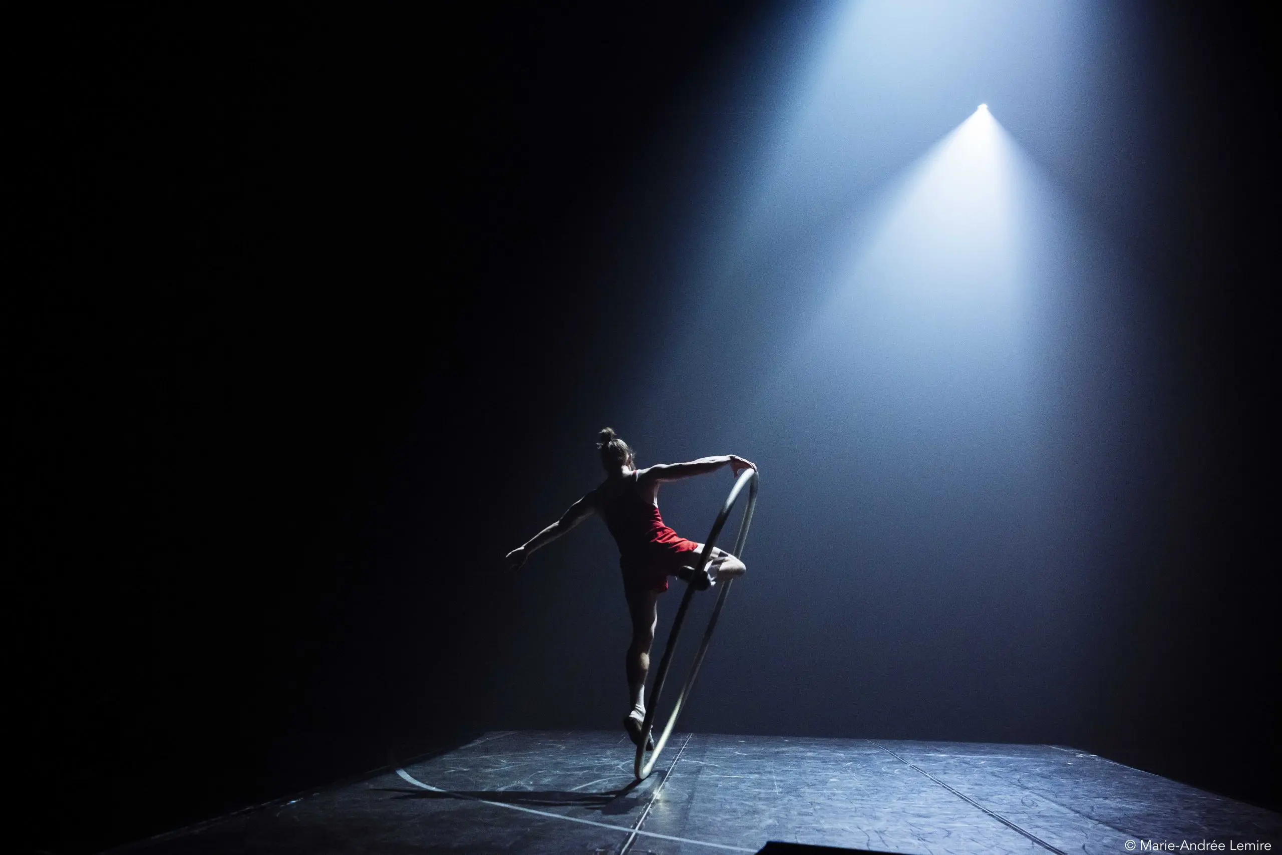 Un artiste se tient gracieusement en équilibre sur une roue Cyr sous un projecteur dramatique sur une scène sombre, sa silhouette évoquant le charisme d'Owen Wilson sur l'arrière-plan aux tons bleus.