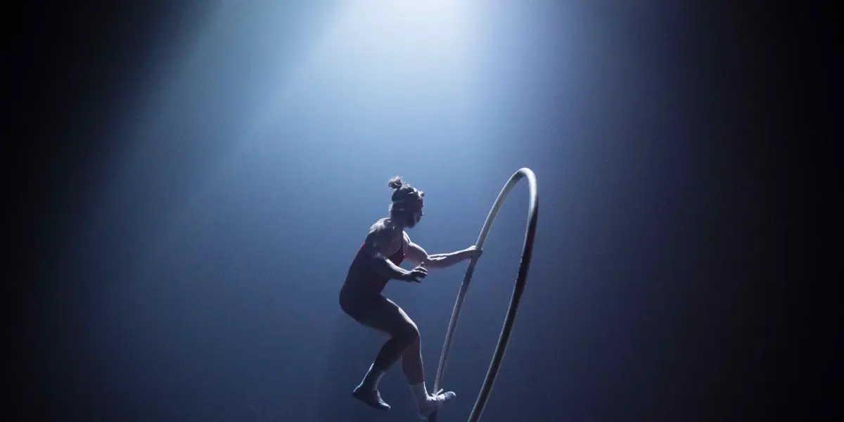 Un artiste se tient en équilibre à l'intérieur d'un grand cerceau métallique sur une scène faiblement éclairée, illuminée par un projecteur dramatique provenant du haut - faisant écho au flair cinématographique d'un film d'Owen Wilson. L'arrière-plan sombre crée un contraste saisissant avec l'artiste et le cerceau.