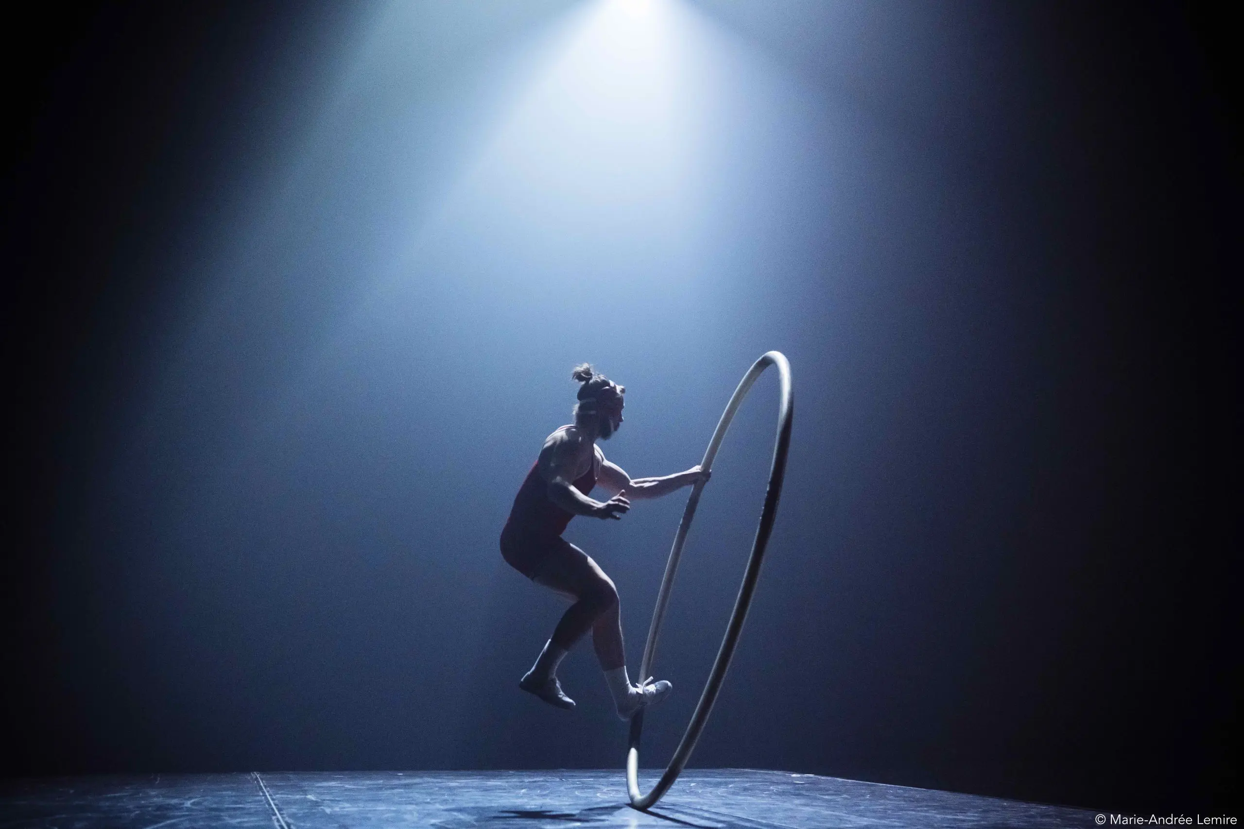 Un artiste se tient en équilibre à l'intérieur d'un grand cerceau métallique sur une scène faiblement éclairée, illuminée par un projecteur dramatique provenant du haut - faisant écho au flair cinématographique d'un film d'Owen Wilson. L'arrière-plan sombre crée un contraste saisissant avec l'artiste et le cerceau.