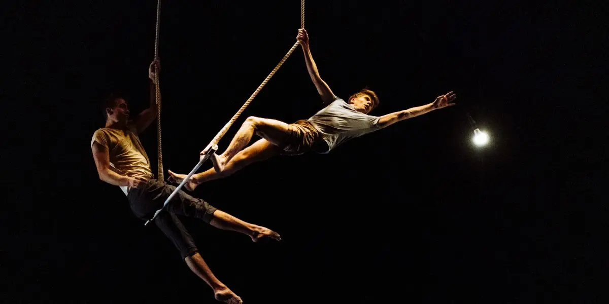 Deux acrobates, Maxime Charron et Corentin Lemaître Auger, évoluent sur des cordes en plein vol sur un fond sombre - l'un s'étirant de manière spectaculaire vers un projecteur lumineux, l'autre offrant son soutien par l'arrière.