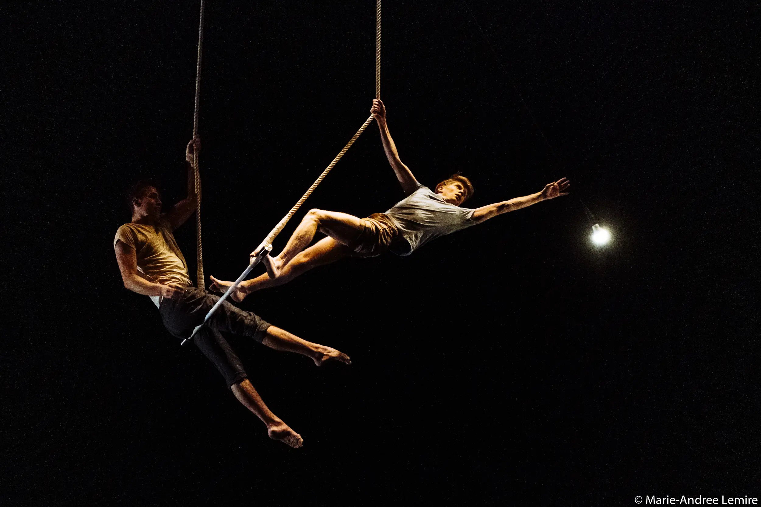 Deux acrobates, Maxime Charron et Corentin Lemaître Auger, évoluent sur des cordes en plein vol sur un fond sombre - l'un s'étirant de manière spectaculaire vers un projecteur lumineux, l'autre offrant son soutien par l'arrière.