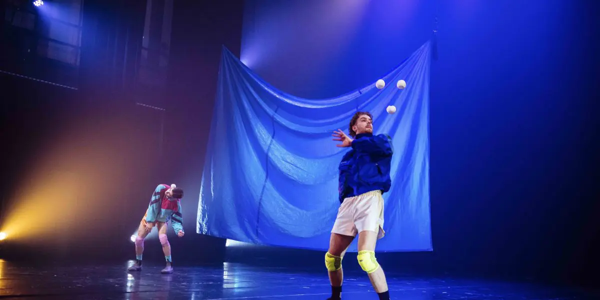 Deux artistes, Zachary Martel et Santiago Rivera Laugerud, sur une scène faiblement éclairée ; l'un jongle avec quatre balles blanches dans des genouillères et une veste bleue tandis que l'autre se tient à l'arrière-plan, partiellement caché par une grande toile de fond bleue.