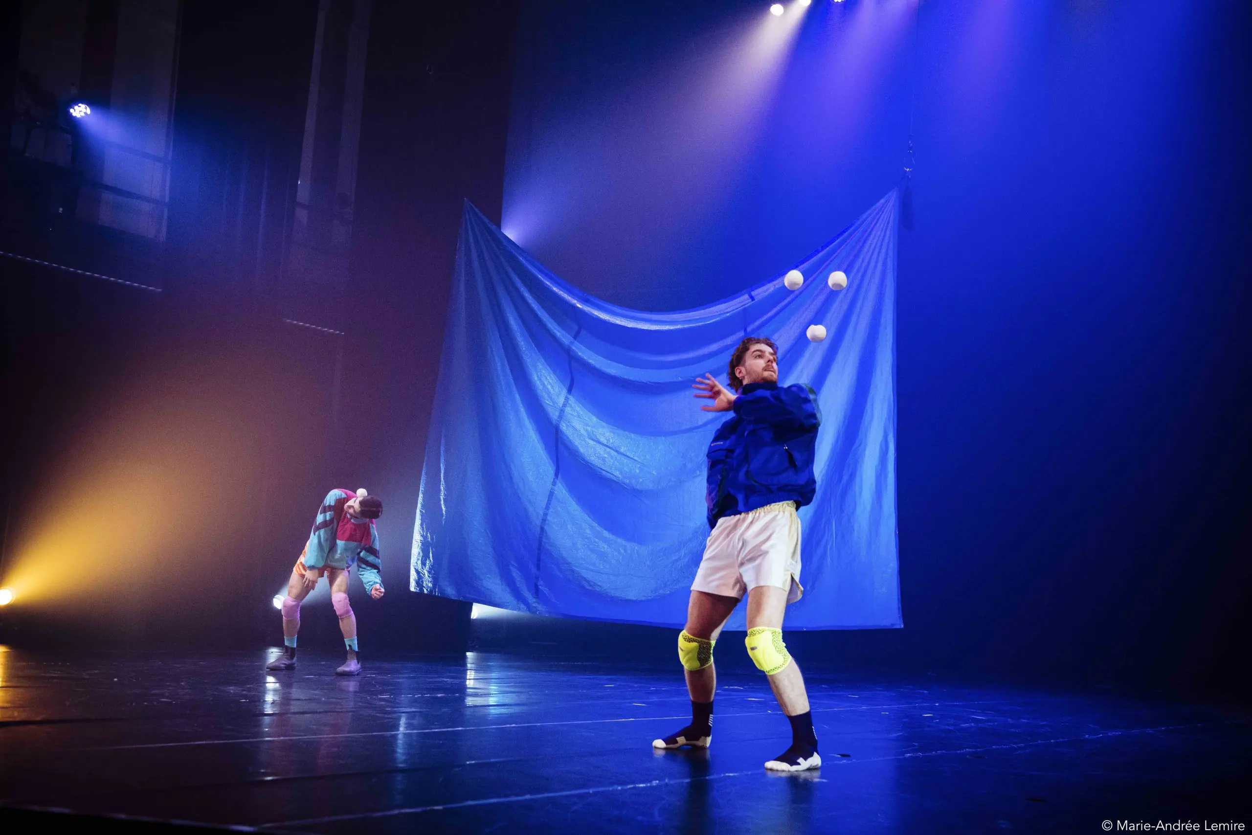 Deux artistes, Zachary Martel et Santiago Rivera Laugerud, sur une scène faiblement éclairée ; l'un jongle avec quatre balles blanches dans des genouillères et une veste bleue tandis que l'autre se tient à l'arrière-plan, partiellement caché par une grande toile de fond bleue.