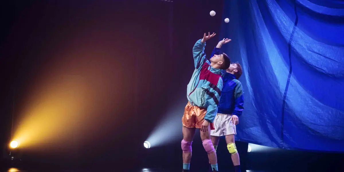 Deux artistes, Zachary Martel et Santiago Rivera Laugerud, vêtus de tenues athlétiques colorées, jonglent avec des balles blanches sur une scène. Un éclairage dramatique et des rideaux bleus plantent le décor alors que l'un d'eux penche la tête en arrière pour équilibrer la balle.